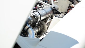 A close-up view of an engine compartment featuring polished metal components, piping, and various mechanical parts. The focus is on the metallic surfaces with bright lighting, highlighting the clean and well-maintained appearance.