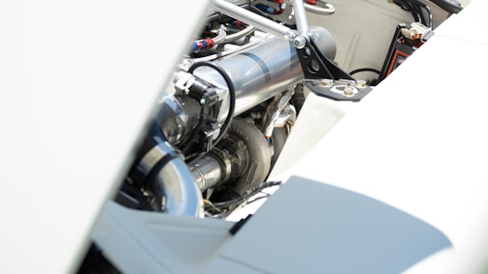 A close-up view of an engine compartment featuring polished metal components, piping, and various mechanical parts. The focus is on the metallic surfaces with bright lighting, highlighting the clean and well-maintained appearance.