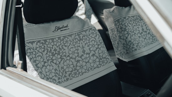 The interior of a car is shown, focusing on the front and rear seats covered with white lace fabric featuring floral patterns. The car is equipped with black headrests, and the lace cover is branded with 'Bluebird' and a website URL.