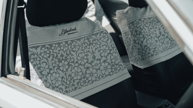 The interior of a car is shown, focusing on the front and rear seats covered with white lace fabric featuring floral patterns. The car is equipped with black headrests, and the lace cover is branded with 'Bluebird' and a website URL.