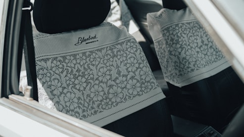 The interior of a car is shown, focusing on the front and rear seats covered with white lace fabric featuring floral patterns. The car is equipped with black headrests, and the lace cover is branded with 'Bluebird' and a website URL.