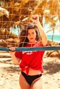 A person with dark, curly hair stands in front of a beach volleyball net, wearing a red top with white text and black bikini bottoms. The background is filled with golden sand and palm trees, suggesting a tropical beach setting.