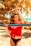A person with dark, curly hair stands in front of a beach volleyball net, wearing a red top with white text and black bikini bottoms. The background is filled with golden sand and palm trees, suggesting a tropical beach setting.