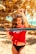 A person with dark, curly hair stands in front of a beach volleyball net, wearing a red top with white text and black bikini bottoms. The background is filled with golden sand and palm trees, suggesting a tropical beach setting.