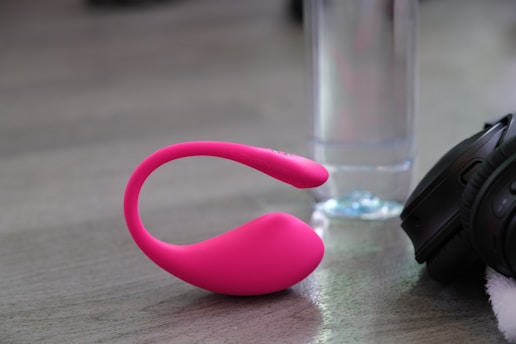 A pink, flexible device rests on a gray surface beside a clear glass or plastic bottle and a pair of black headphones. The device is sleek and modern in design, with a smooth texture.