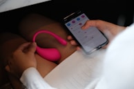 A person is holding a smartphone with an app interface visible on the screen. The other hand holds a pink, curvy object with a smooth texture. The setting appears to be indoors, possibly in a private or relaxed environment.