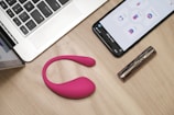 On a wooden table, there is an open laptop, a smartphone displaying an app interface, a pink wearable device, and a metallic cylindrical object, possibly a cosmetic tube.