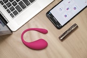 On a wooden table, there is an open laptop, a smartphone displaying an app interface, a pink wearable device, and a metallic cylindrical object, possibly a cosmetic tube.