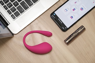 On a wooden table, there is an open laptop, a smartphone displaying an app interface, a pink wearable device, and a metallic cylindrical object, possibly a cosmetic tube.