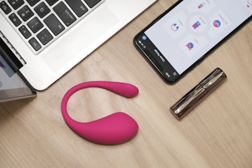 On a wooden table, there is an open laptop, a smartphone displaying an app interface, a pink wearable device, and a metallic cylindrical object, possibly a cosmetic tube.