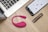 On a wooden table, there is an open laptop, a smartphone displaying an app interface, a pink wearable device, and a metallic cylindrical object, possibly a cosmetic tube.