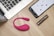 On a wooden table, there is an open laptop, a smartphone displaying an app interface, a pink wearable device, and a metallic cylindrical object, possibly a cosmetic tube.