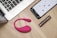 On a wooden table, there is an open laptop, a smartphone displaying an app interface, a pink wearable device, and a metallic cylindrical object, possibly a cosmetic tube.