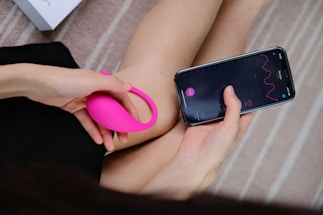 A person holds a bright pink wearable device in one hand while interacting with a smartphone in the other hand. The smartphone screen displays a graph with lines and numerical data, suggesting a tracking or monitoring application. The background consists of a soft, neutral-toned surface with a partial view of a box.