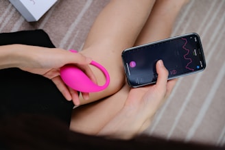 A person holds a bright pink wearable device in one hand while interacting with a smartphone in the other hand. The smartphone screen displays a graph with lines and numerical data, suggesting a tracking or monitoring application. The background consists of a soft, neutral-toned surface with a partial view of a box.