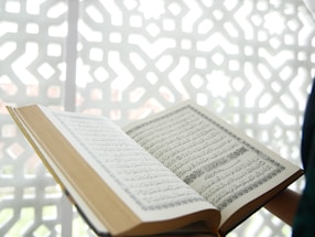 An open book featuring Arabic script is displayed prominently, with a geometric, patterned background in white. The book appears to be the focal point, with its pages spread and text clearly visible.
