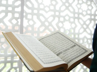 An open book featuring Arabic script is displayed prominently, with a geometric, patterned background in white. The book appears to be the focal point, with its pages spread and text clearly visible.