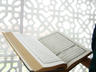 An open book featuring Arabic script is displayed prominently, with a geometric, patterned background in white. The book appears to be the focal point, with its pages spread and text clearly visible.