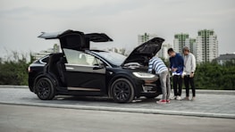 Three people stand by a black car with both the front trunk and rear passenger doors open, examining the front area. The scene takes place on a paved surface with greenery and tall buildings in the background, suggesting an urban setting.