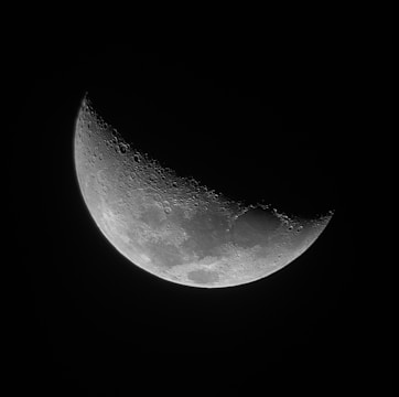 A crescent moon with detailed craters and surface textures against a dark, black background. The lunar surface appears intricate and textured, highlighting the contrast between the illuminated and shadowed parts.