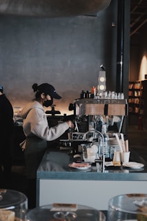 A barista in a white hoodie and dark cap is operating an espresso machine in a modern coffee shop setting. Various items such as cups, napkins, and a water bottle are neatly arranged on the counter. The background shows shelves filled with items, giving a cozy and minimalistic vibe to the interior.