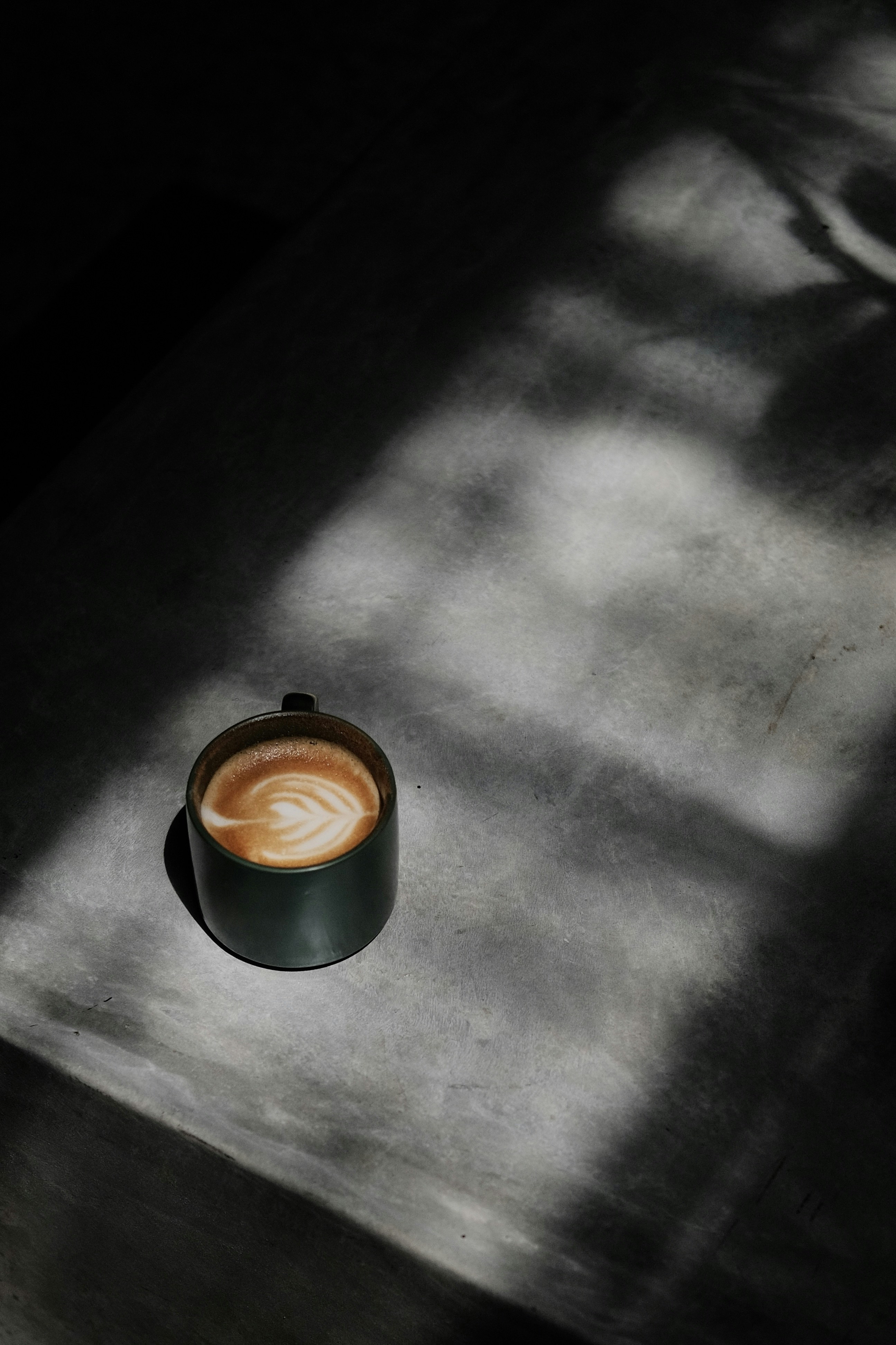 A single dark mug of latte sits on a textured concrete surface, bathed in diagonal light that creates dramatic shadows.