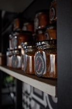 A collection of glass jars containing a reddish-brown substance, likely a type of sauce or spread, is neatly arranged on a wooden shelf. The jars have black lids and labels with red and white text and graphics, including a red chili pepper. The setting appears to be a dimly lit pantry or storage space with a focus on the organization and presentation of the jars.