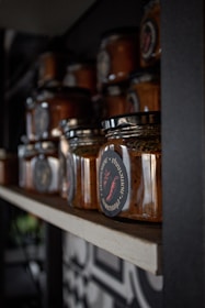 A collection of glass jars containing a reddish-brown substance, likely a type of sauce or spread, is neatly arranged on a wooden shelf. The jars have black lids and labels with red and white text and graphics, including a red chili pepper. The setting appears to be a dimly lit pantry or storage space with a focus on the organization and presentation of the jars.