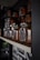 A collection of glass jars containing a reddish-brown substance, likely a type of sauce or spread, is neatly arranged on a wooden shelf. The jars have black lids and labels with red and white text and graphics, including a red chili pepper. The setting appears to be a dimly lit pantry or storage space with a focus on the organization and presentation of the jars.
