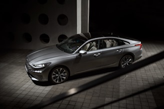 A sleek, modern sedan with metallic silver paint parked indoors on a textured tile floor. The car features sharp lines and a stylish design, with contemporary alloy wheels and distinctive front and rear lights. The background is a dark space with round cutouts visible on a cement wall.