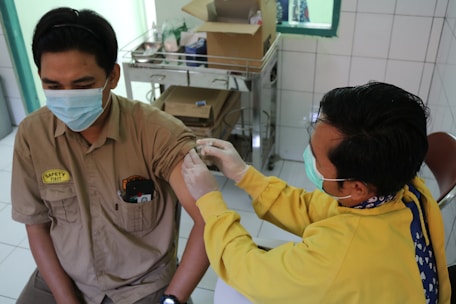 A person wearing a medical mask receives an injection in the upper arm from a healthcare worker in a yellow uniform. The setting appears to be a medical or clinical environment with tiled walls and a cart with supplies in the background.