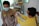 A person wearing a medical mask receives an injection in the upper arm from a healthcare worker in a yellow uniform. The setting appears to be a medical or clinical environment with tiled walls and a cart with supplies in the background.