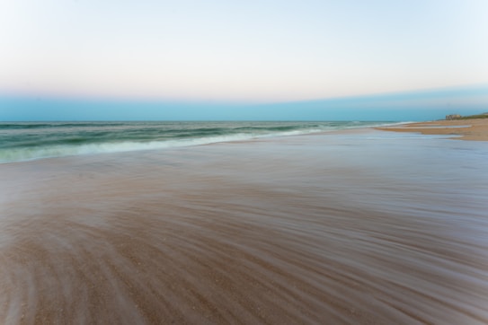 A serene coastal scene featuring gentle ocean waves lapping against a deserted sandy beach. The sky is a gradient of soft pastels, with hints of pink and blue blending harmoniously. Minimalistic and calming, the horizon is a seamless blend where sea meets sky.