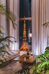 A cozy, modern living room with large windows displaying an illuminated city tower at night. The room features a white sofa, a wooden coffee table with candles, decorative plants, and light-colored curtains. The vibrant orange and yellow lights of the tower contrast with the calm and neutral tones of the room's decor.