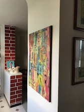 A vibrant, abstract painting featuring multiple faces and eyes with a variety of colors on a wall in a well-lit room. The wall is adjacent to a brick-patterned section, and there are additional decorations, such as a small framed artwork and flowers, on a white ledge.