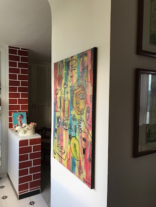 A vibrant, abstract painting featuring multiple faces and eyes with a variety of colors on a wall in a well-lit room. The wall is adjacent to a brick-patterned section, and there are additional decorations, such as a small framed artwork and flowers, on a white ledge.