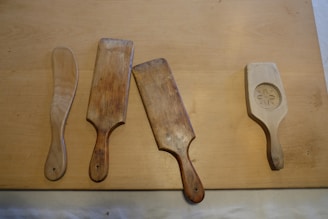 Four wooden spatulas are arranged on a flat wooden surface. Each spatula has a different shape and patina, showing variation in age and use. The far right spatula features a carved flower design.