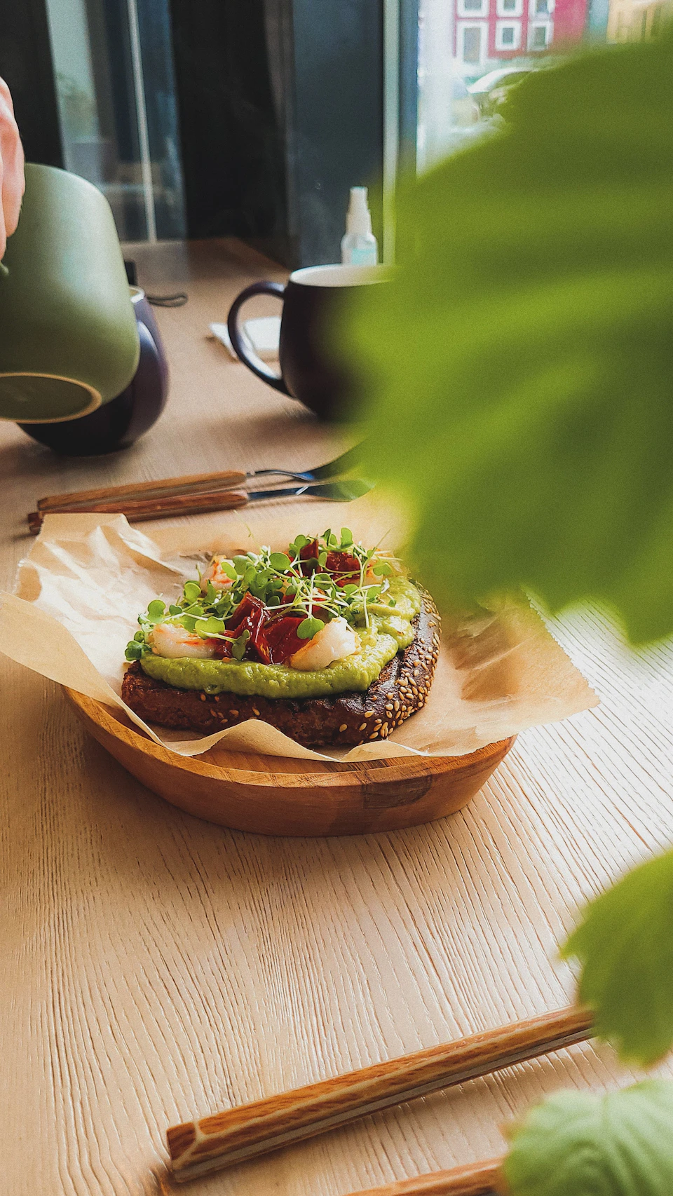 The image shows a wooden table with various objects on it, including a plate holding an avocado toast sandwich and a cup filled with coffee. The avocado toast is topped with red onions and other toppings. A pair of chopsticks rests next to the plate, suggesting that the meal may be enjoyed using these utensils. In the background, there are two potted plants - one on each side of the table.