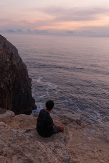 A serene solo female traveler sitting on a rocky cliff overlooking a calm sea at sunset.