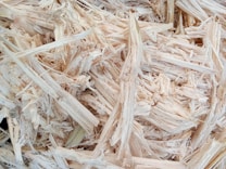 A close-up view of light-colored wood shavings or chips, showcasing the fibrous and textured nature of the material. The shavings appear dry and randomly stacked with varying widths and lengths.