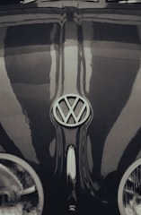 A vintage Volkswagen Beetle with a prominently displayed emblem on the hood, exhibiting classic curves and a glossy finish. The image has a retro, monochrome style with reflections visible on the surface of the car.