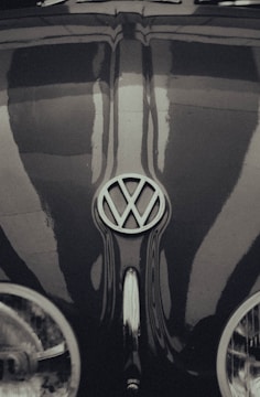 A vintage Volkswagen Beetle with a prominently displayed emblem on the hood, exhibiting classic curves and a glossy finish. The image has a retro, monochrome style with reflections visible on the surface of the car.