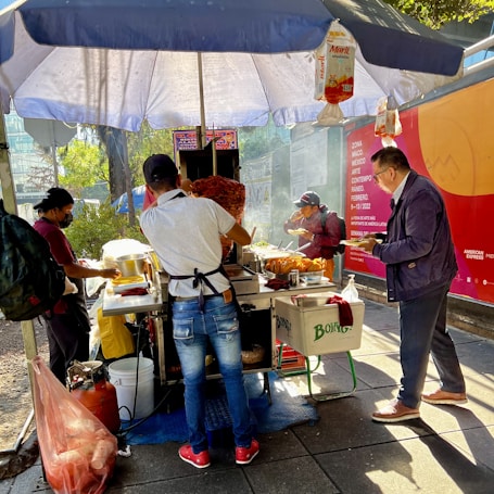 A street food vendor serves tacos under a large umbrella. Several people are gathered around the stand, with one man cooking and others eating. The setup includes a vertical rotisserie with meat, and various condiments and supplies are visible. There is a bright, sunny atmosphere with dappled shadows on the ground.