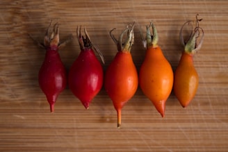 Five small, vibrant fruits in shades of red and orange are arranged in a line on a wooden surface. The fruits have an elongated shape with dried, brownish stems at the top.