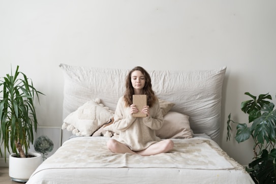 A person sits cross-legged on a bed, holding a book. The setting is serene, with neutral colors and soft textures. Plants are placed on either side, adding a touch of greenery to the minimalist decor.