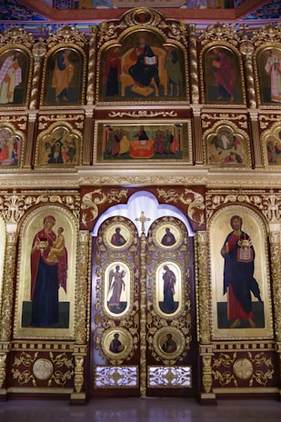 A richly adorned iconostasis featuring multiple religious icons set in ornate golden frames. The central doors, known as the Royal Doors, are intricately designed with religious imagery. The upper panels depict various saints and religious figures, while the surrounding architecture includes elaborate decorative elements, showcasing a blend of red, gold, and other vibrant colors.