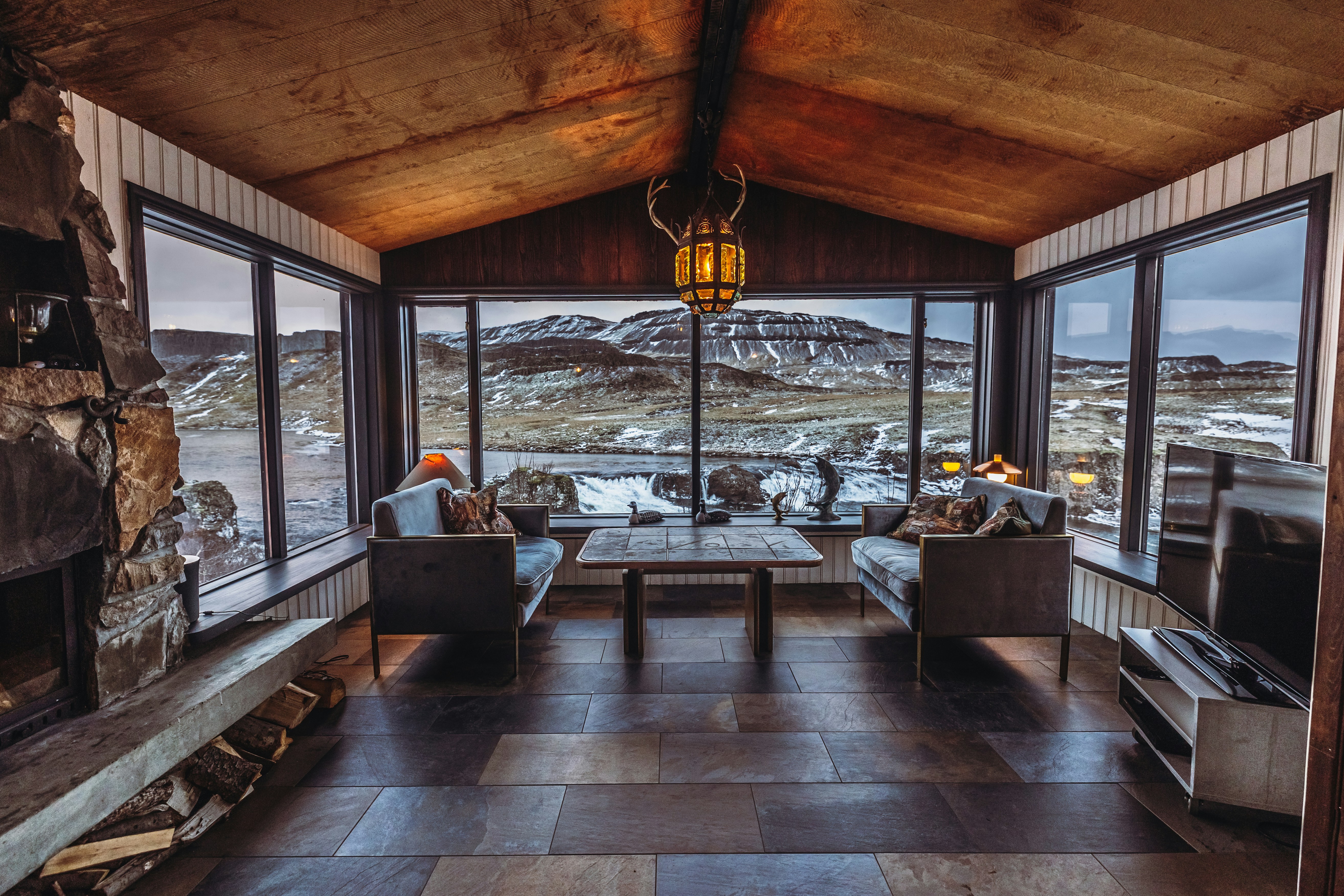 A cozy room with large glass windows offering a panoramic view of a snowy landscape and hills in the distance. The room includes two modern sofas facing each other with a wooden table between them. A stone fireplace is on one side with chopped wood neatly stacked below. A warm, rustic chandelier hangs from the wooden ceiling, adding to the inviting atmosphere.