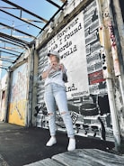 A person stands in front of a mural featuring typewriters and the text 'I'll forever wish I found words I would write.' The scene is set in an industrial-looking area with exposed structural beams and graffiti art nearby. The person is wearing casual clothing, including a cap, a graphic t-shirt, ripped jeans, and sneakers.