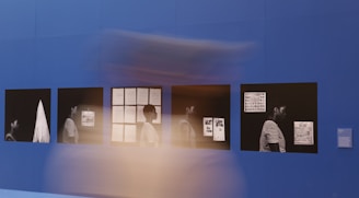 The image displays a series of framed photographs or artworks on a deep blue wall. These frames contain monochromatic images, each featuring a person in a striped shirt, shown in various poses with different documents in the background. There is a blur of motion in front of these frames, suggesting movement.