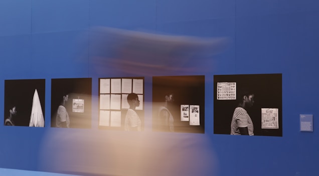 The image displays a series of framed photographs or artworks on a deep blue wall. These frames contain monochromatic images, each featuring a person in a striped shirt, shown in various poses with different documents in the background. There is a blur of motion in front of these frames, suggesting movement.